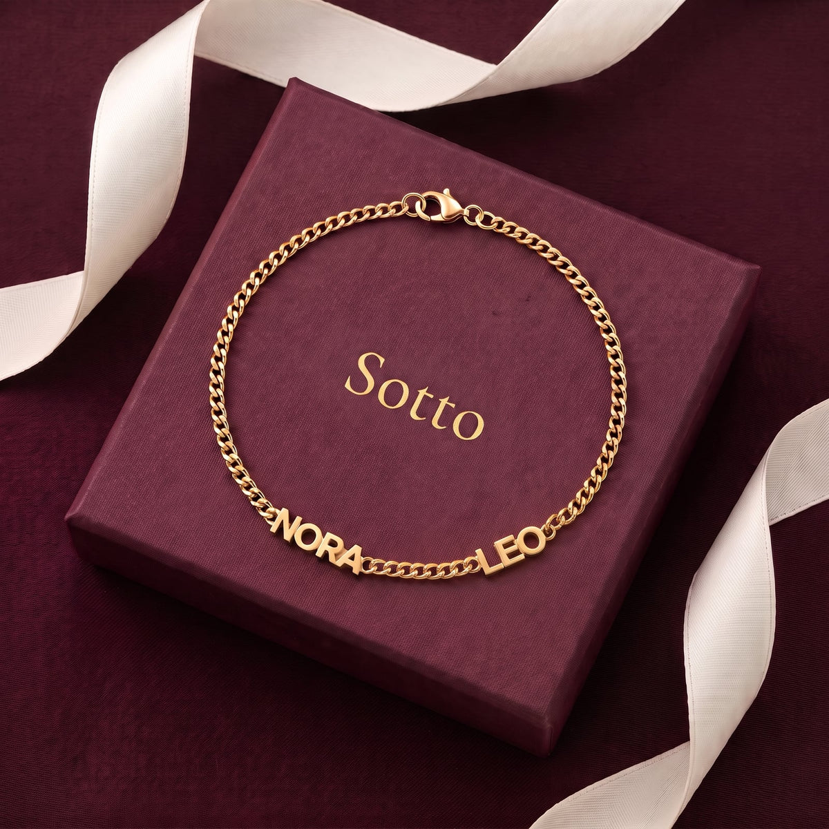 Gold NORA and LEO name bracelet laid on a wine-red Sotto gift box with ivory satin ribbon