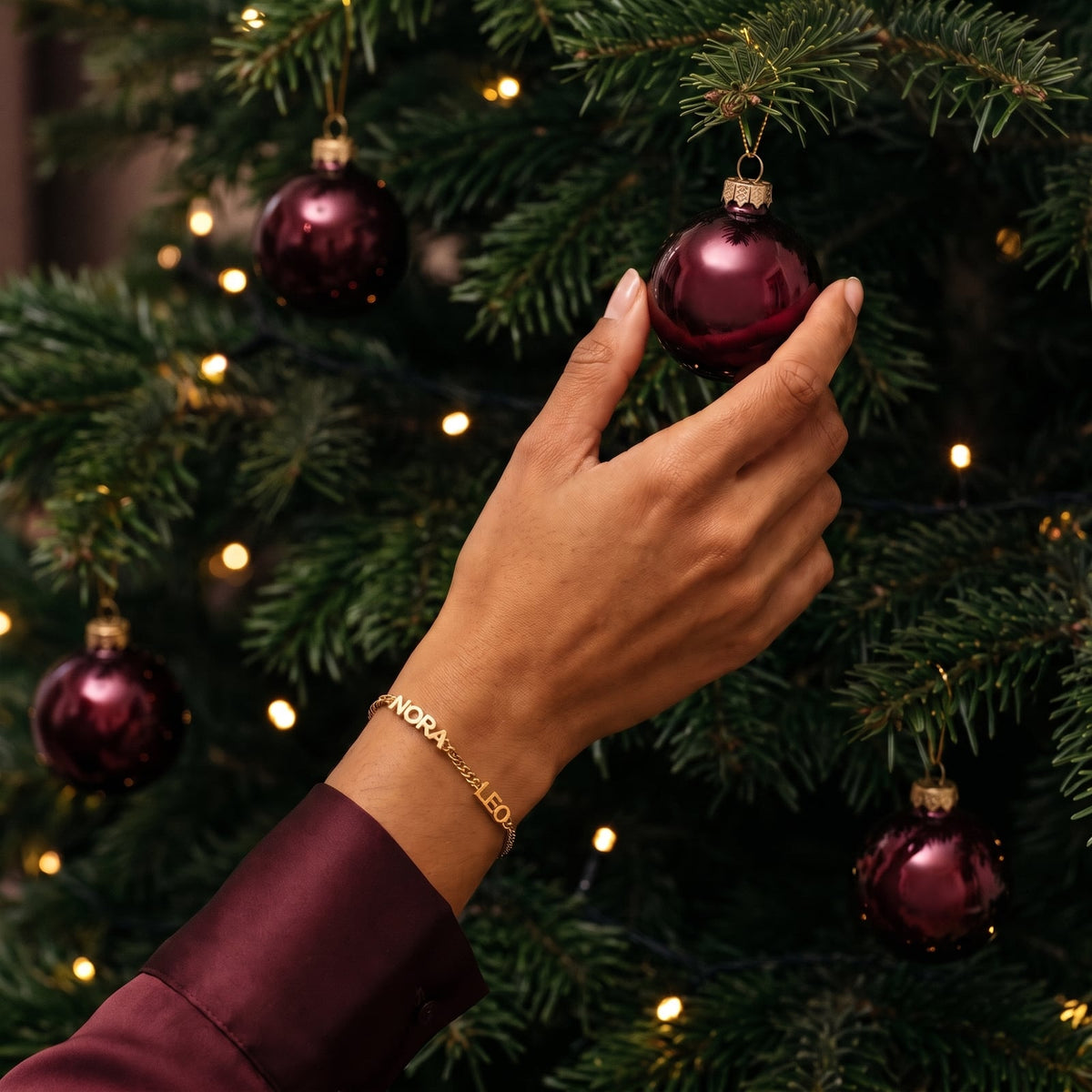 Gold personalised name bracelet reading NORA and LEO worn on a wrist, hand hanging a wine-red bauble on a Christmas tree